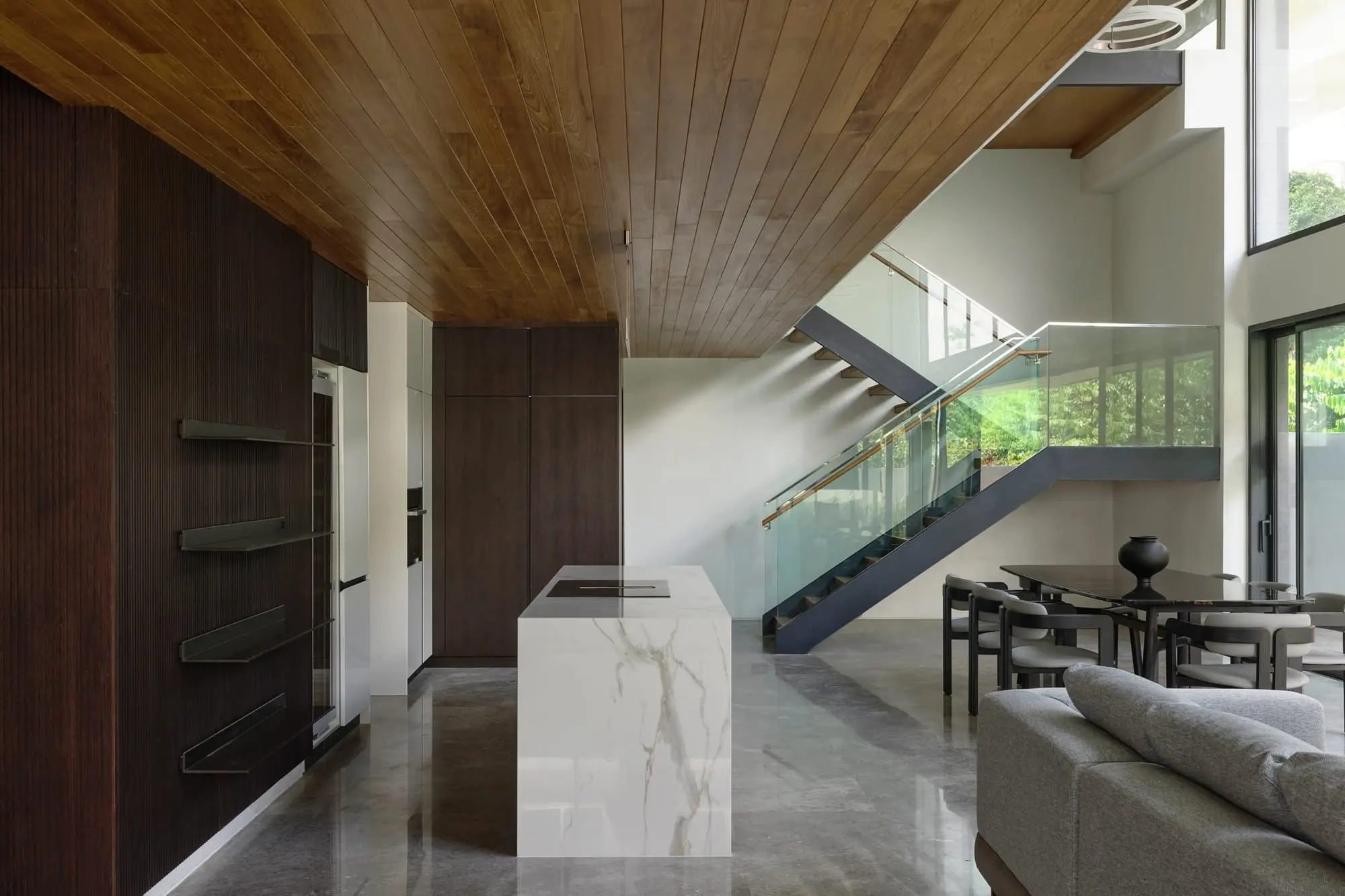 Living and kitchen space under a timber ceiling, with a marble island and a steel and glass stair beyond